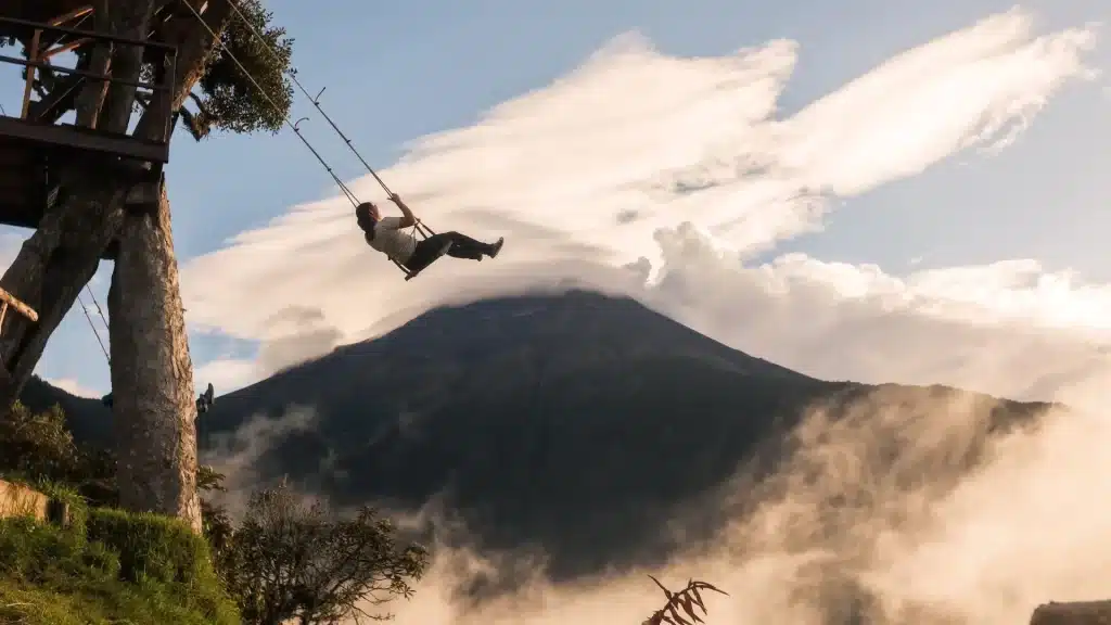A person swinging over a cliff at the Swing at the End of the World near La Casa del Árbol in Baños, Ecuador, with Tungurahua Volcano in the background.