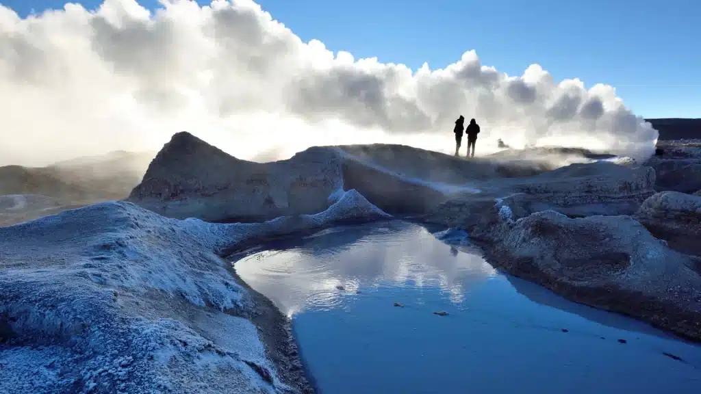 Silhouettes of two people walking near a steaming hot spring in the Bolivian Andes, with a shallow lake reflecting the soft light.