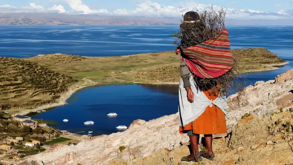Aymara or Quechua woman in traditional clothing near the shores of Lake Titicaca, with the lake and Andes in the background.
