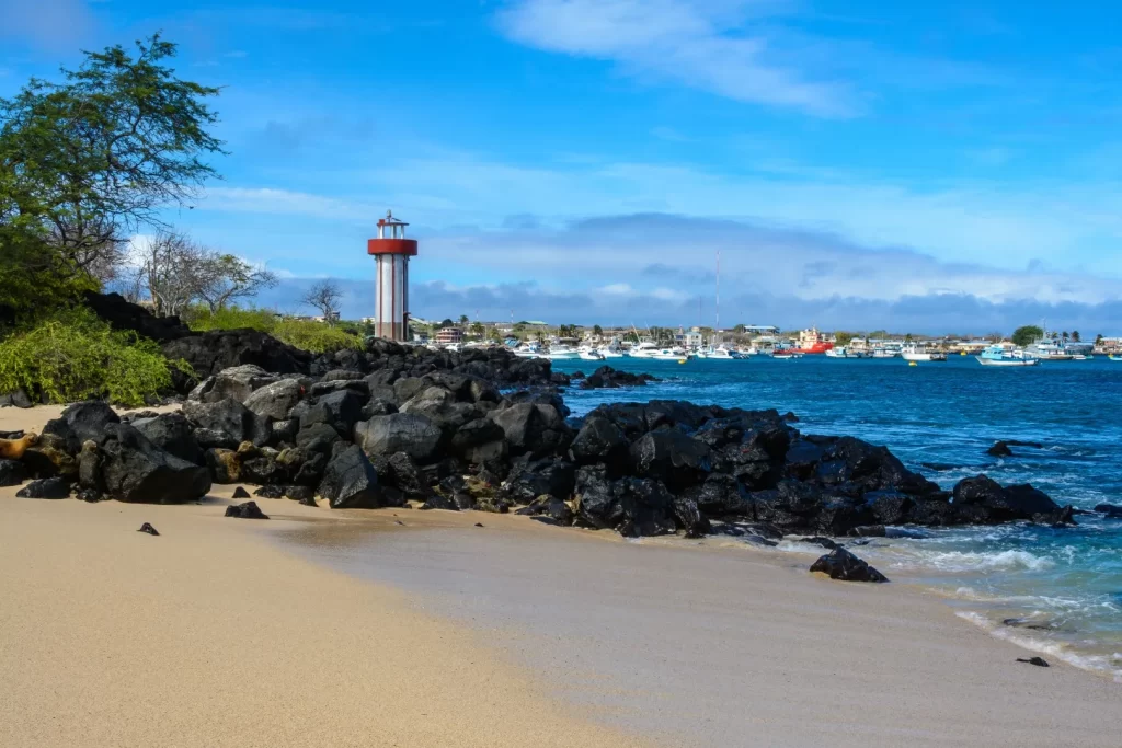 Rocky beach with black volcanic stones, a lighthouse, and boats in the distance on San Cristóbal Island, Galápagos. - Darwin islands visited