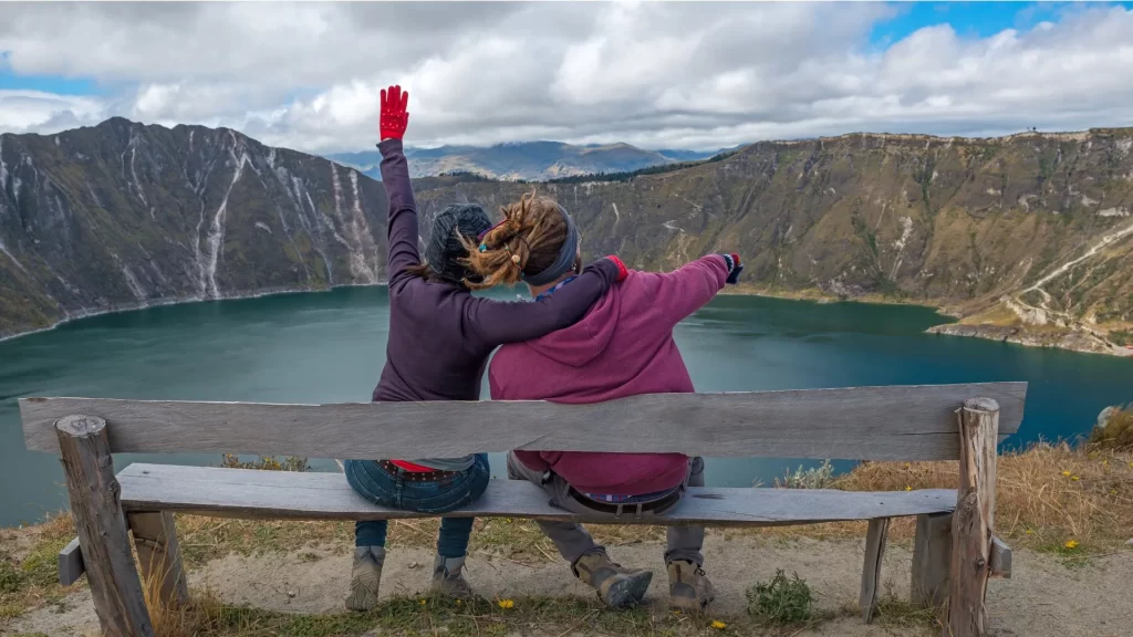 The image shows two people sitting on a wooden bench, overlooking the Quilotoa Lagoon in Ecuador-Trekking in Ecuador