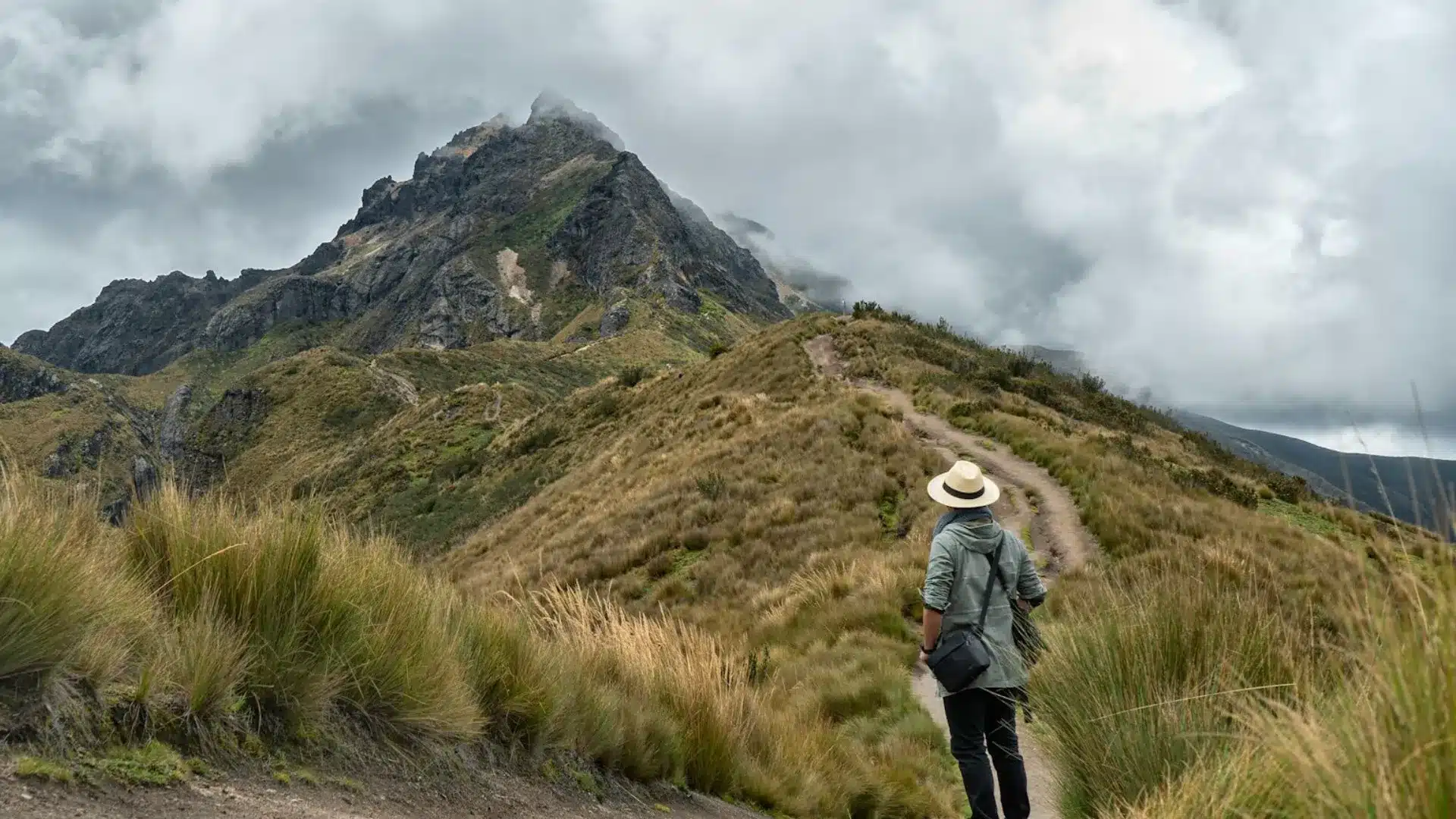Hiker with hat and backpack walking along a dirt path in the páramo toward Pichincha Volcano under a cloudy sky.